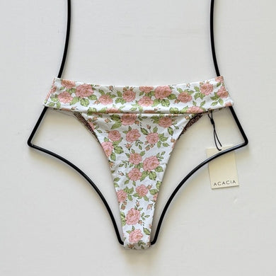 Flat lay of Acacia Swimwear Mateo Bikini Bottom in Rosie floral print, featuring blush pink roses on a white background with green leaves.
