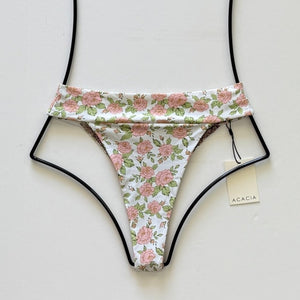 Flat lay of Acacia Swimwear Mateo Bikini Bottom in Rosie floral print, featuring blush pink roses on a white background with green leaves.
