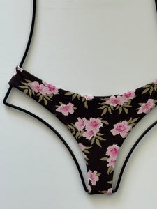 Close-up of Acacia Ho’okipa bikini bottom in Floret print. Dark brown background with pink flowers and olive leaves, low-rise cheeky fit.