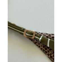 Close-up of Acacia Swimwear Ralphy Bikini Bottom in Hinahina, showing matte logo hardware engraved with “ACACIA” and adjustable side strap detail over ribbed tan fabric with subtle animal-print lining beneath.