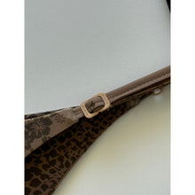 Close-up of Acacia Swimwear Ralphy Bikini Bottom in Aimi, showing matte logo hardware engraved with “ACACIA” and adjustable side strap detail over ribbed tan fabric with subtle animal-print lining beneath.